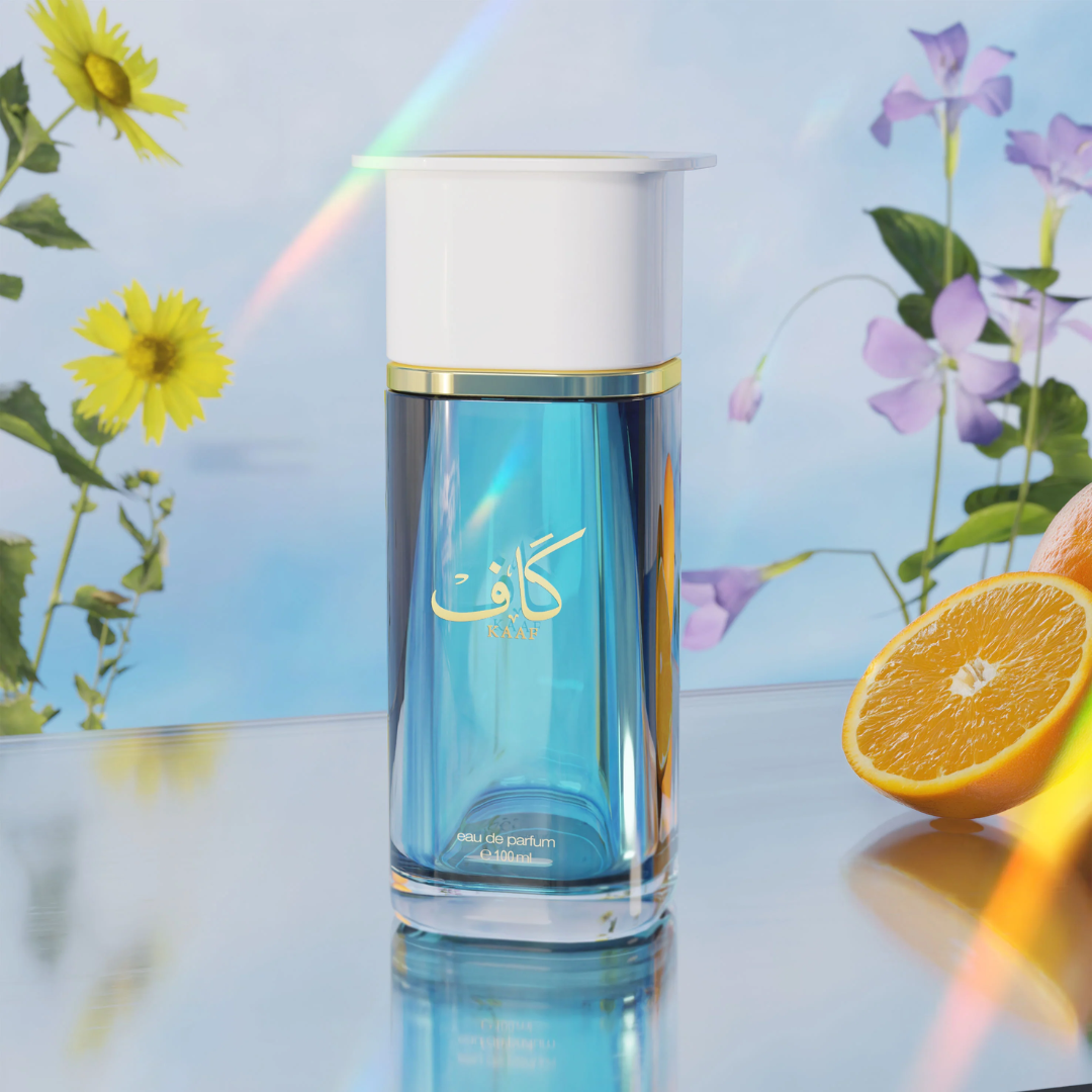 Blue perfume bottle with white cap on a reflective surface with flowers and an orange in the background. Ahmed Al Maghribi Kaaf Unisex Perfume