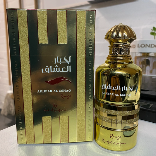 Gold perfume bottle and packaging with 'Akhbar Al Ushaq' branding on a reflective surface. Akhbar Al Ushaq Rouge by Ard Al Zaafaran Womens Perfume