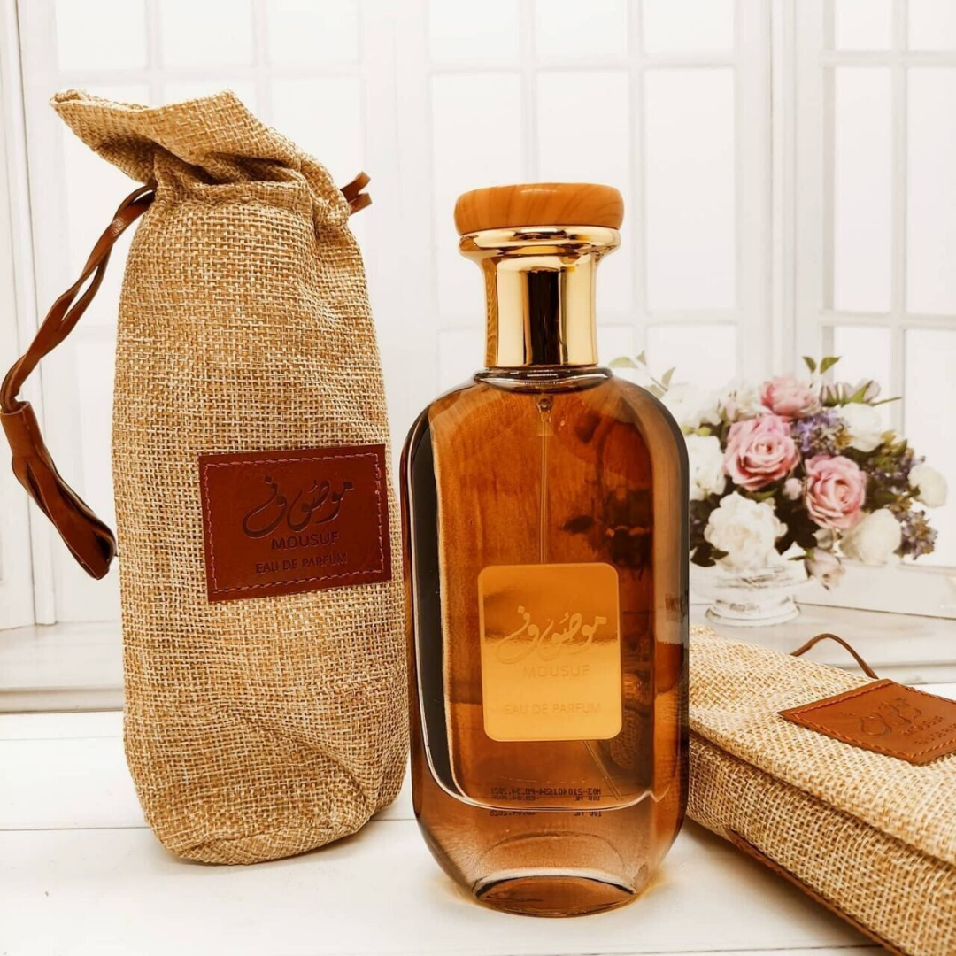 Mousuf Perfume Display with Flowers – Luxury Mousuf Eau de Parfum bottle alongside its signature burlap pouch, set in a bright floral backdrop for an opulent and romantic aesthetic. A glass bottle of Mousuf Perfume by Ard Al Zaafaran with a brown liquid inside, placed next to its burlap fabric packaging with a leather patch and a brown label. 