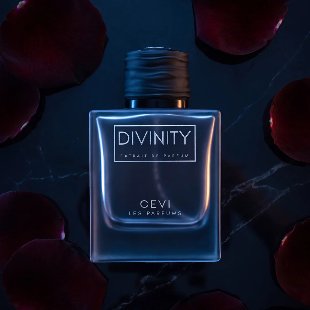 A square glass bottle of Divinity Extrait de Parfum by Cevi Les Parfums, featuring a dark metallic cap and a blue-toned label, set against a dark background with scattered red rose petals.