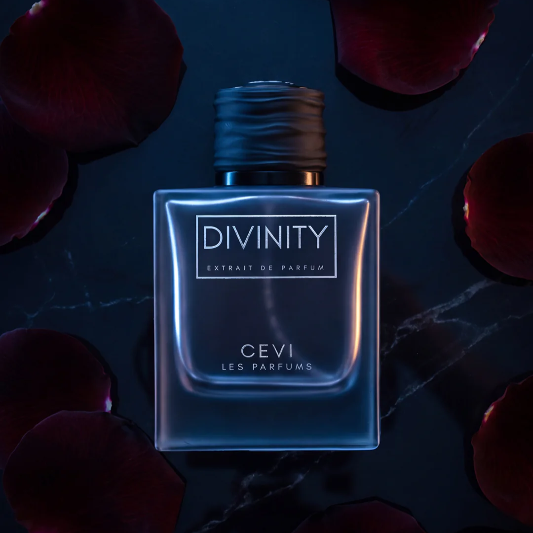 A square glass bottle of Divinity Extrait de Parfum by Cevi Les Parfums, featuring a dark metallic cap and a blue-toned label, set against a dark background with scattered red rose petals.