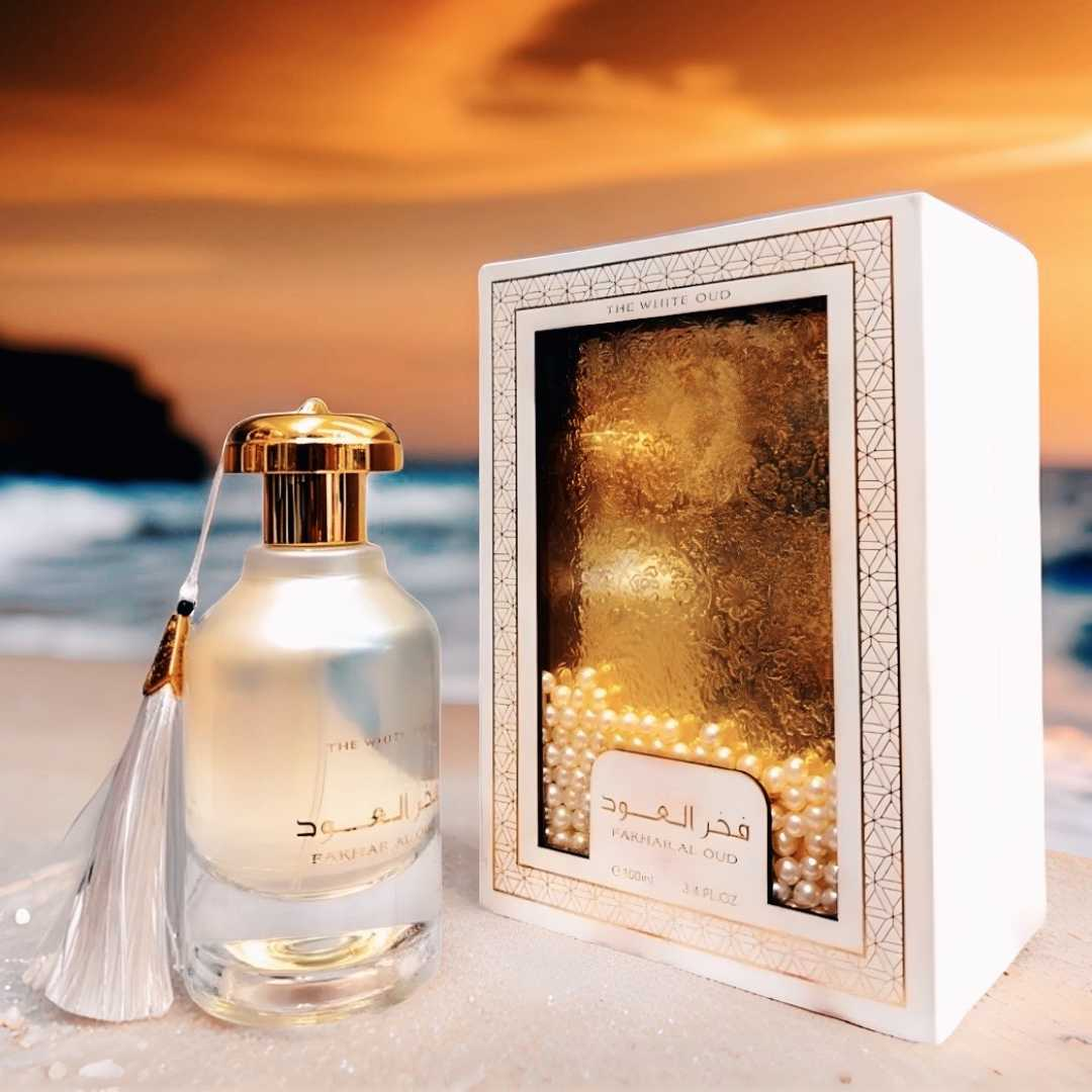 Perfume bottle with gold cap and tassel next to its packaging on a beach at sunset. Fakhar Al Oud The White Oud by Ard Al Zaafaran Unisex Perfume