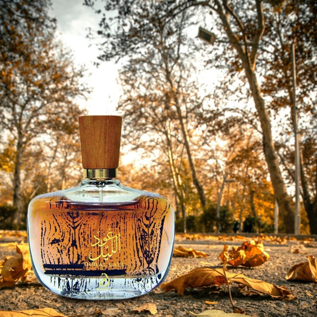 Perfume bottle with wooden cap on a leaf-covered ground with trees in the background. Oud Al Layl by Arabiyat Unisex Perfume