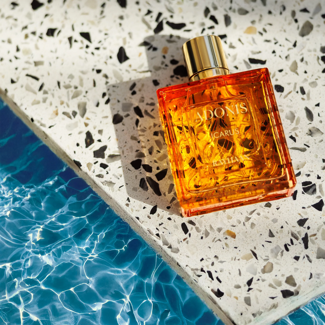Orange perfume bottle with gold cap on a textured surface near water. Rayhaan Adonis Icarus Mens Perfume