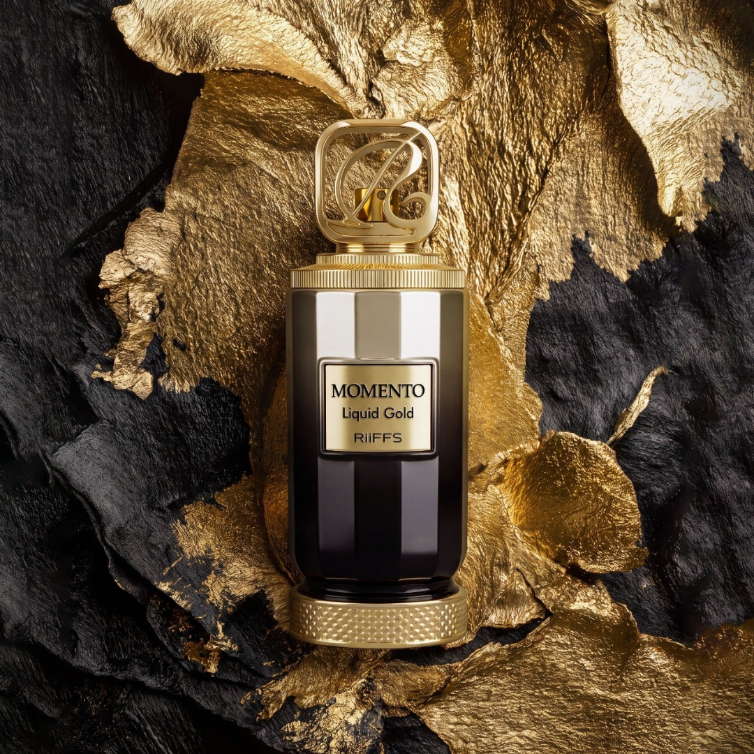 A luxury perfume bottle labeled 'MOMENTO Liquid Gold RiiFFS' sits centered on a dramatic background of textured gold and black leaves. The bottle features a dark, glossy body with a metallic gold cap and base, and a gold label on the front.