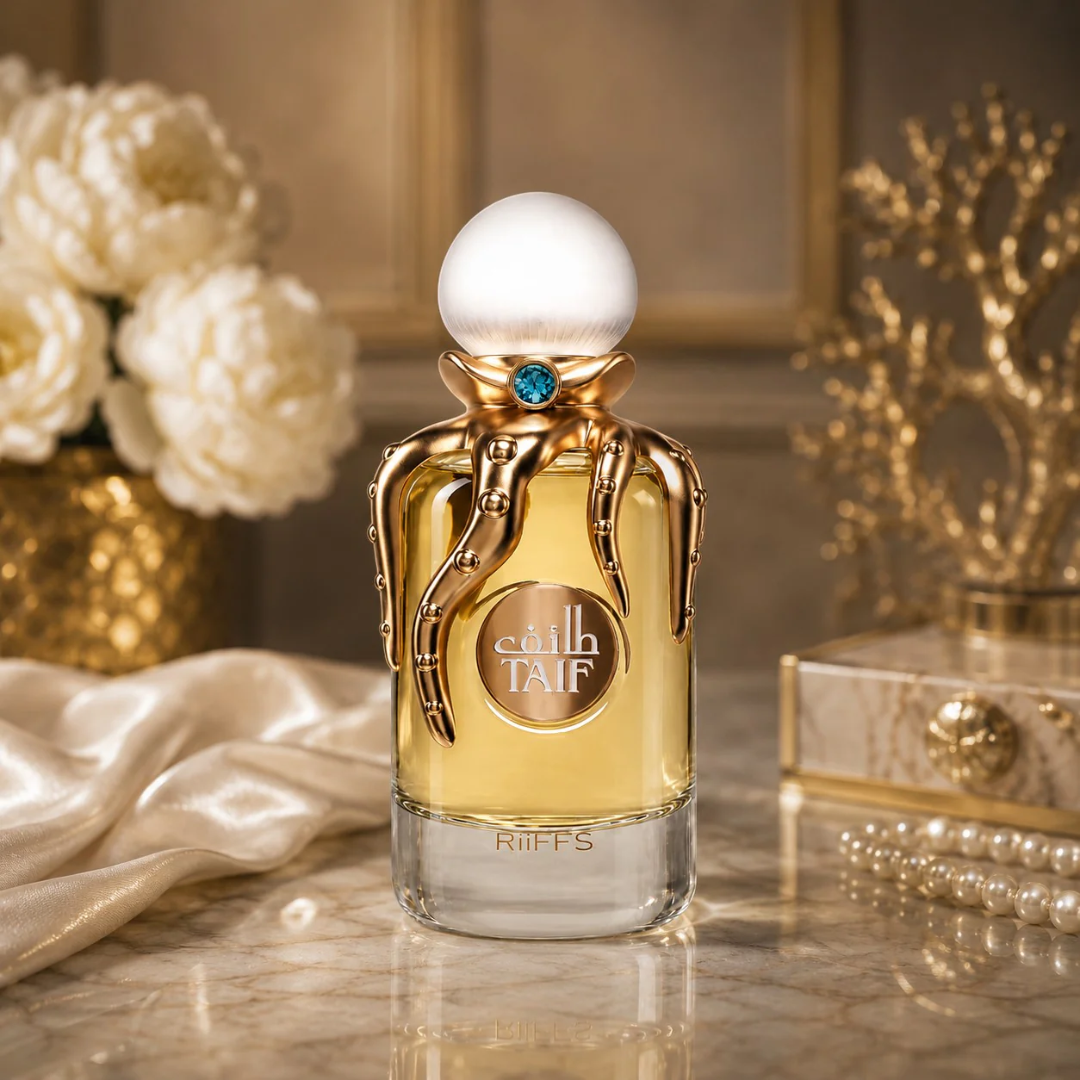 A bottle of Taif RIFFS perfume stands on a marble surface in a luxurious interior setting. The bottle has a gold cap with a white spherical top, a blue gemstone, and gold tent-like details wrapping around the neck.