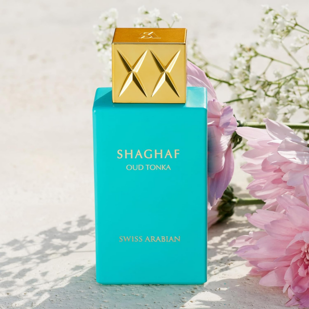Turquoise perfume bottle with gold cap labeled 'Shaghaf Oud Tonka' by Swiss Arabian, surrounded by pink flowers.