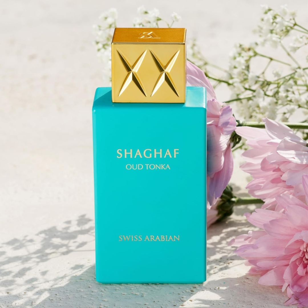 Turquoise perfume bottle with gold cap labeled 'Shaghaf Oud Tonka' by Swiss Arabian, surrounded by pink flowers.