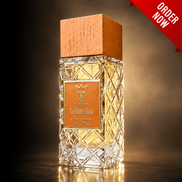 A faceted crystal perfume bottle with a natural wood cap and an orange label reading 'Bujairami Too Damn Good', shown on a dark reflective surface with a warm spotlight. A red 'ORDER NOW' banner appears in the top right corner.